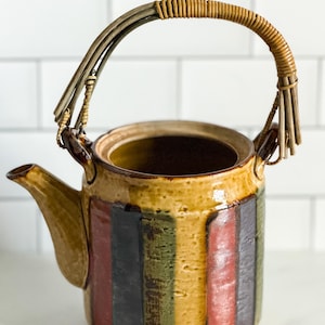 May include: A ceramic teapot featuring a cylindrical body with vertical stripes in shades of mustard yellow, dark green, and rust red. The teapot has a curved spout and a tall, arched handle wrapped in thin, light brown natural fiber material.