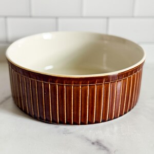 May include: A round ceramic baking dish featuring a deep brown exterior with a vertical ribbed texture and a smooth, cream-coloured interior. The vessel has a wide, open top and a flat base, designed for culinary use in a kitchen or dining setting.