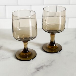 May include: Two matching wine glasses crafted from translucent smoke-colored glass. Each glass features a rounded bowl, a short, sturdy stem, and a circular base. The glass has a smooth, uniform finish with a consistent dark brown tint throughout.