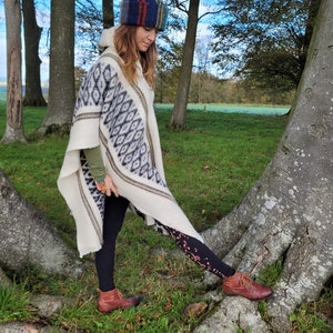 May include: A woman wearing a white poncho with a black and white geometric pattern, a colorful knitted hat, black leggings with a musical note pattern, and brown boots. She is standing in a forest setting.