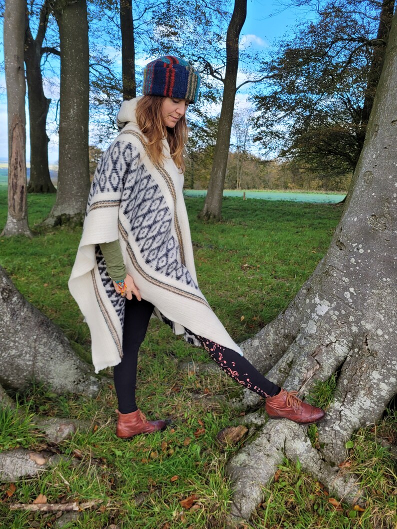 May include: A woman wearing a white poncho with a black and white geometric pattern, a colorful knitted hat, black leggings with a musical note pattern, and brown boots. She is standing in a forest setting.