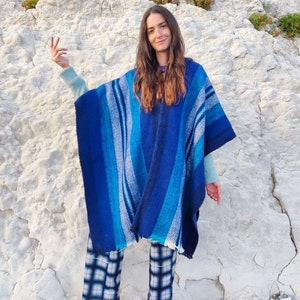 May include: A woman wearing a blue and white striped poncho with a fringe hem. She is standing in front of a white rocky cliff and wearing black and white plaid pants.