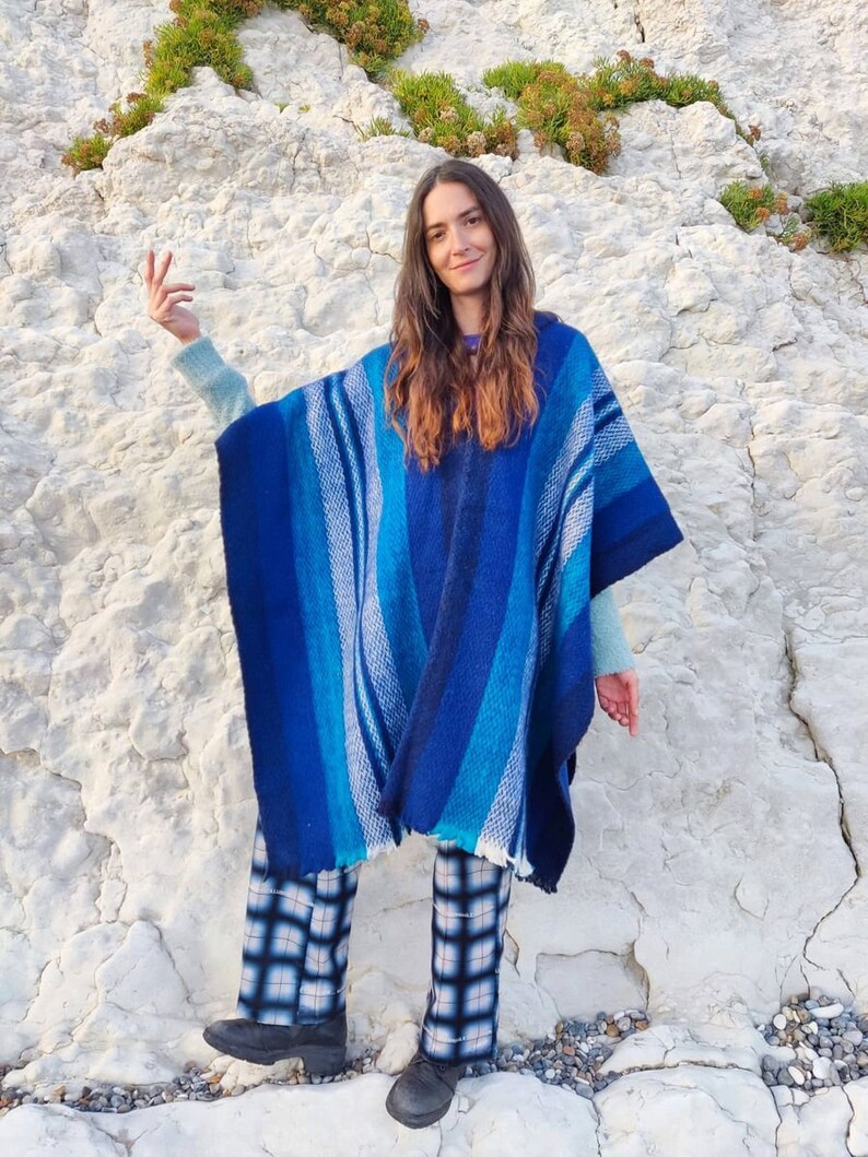 May include: A woman wearing a blue and white striped poncho with a fringe hem. She is standing in front of a white rocky cliff and wearing black and white plaid pants.