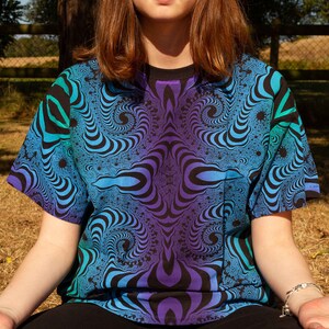 May include: A person wearing a blue, green and purple t-shirt with a swirling, psychedelic pattern. The person is sitting with their legs crossed and their eyes closed.