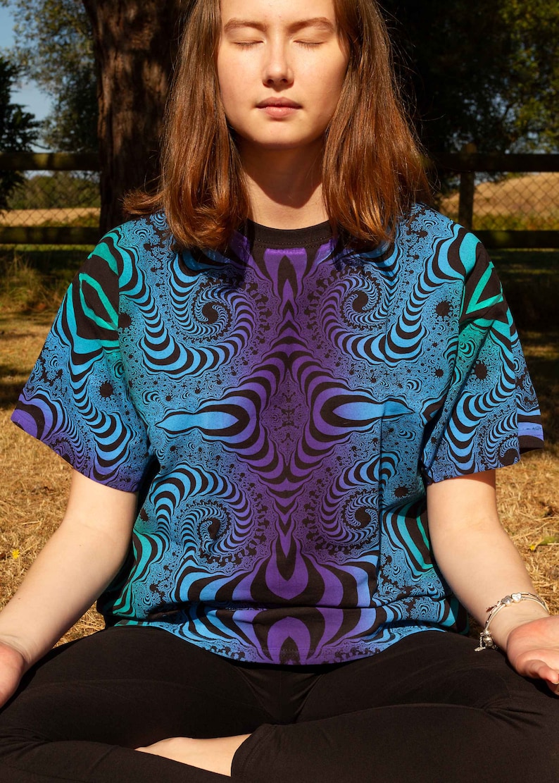 May include: A person wearing a blue, green and purple t-shirt with a swirling, psychedelic pattern. The person is sitting with their legs crossed and their eyes closed.