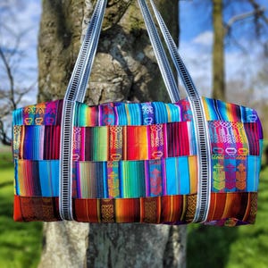 May include: A colourful, woven duffel bag with white straps. The bag is made of a patchwork of different coloured fabrics, including red, blue, green, yellow, and purple.