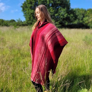 May include: A woman wearing a red hooded poncho with a striped pattern, standing in a field of tall grass.