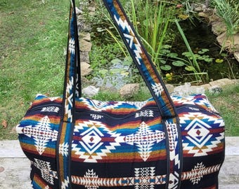 Puede incluir: Una bolsa de viaje con base negra y un vibrante patrón geométrico en tonos azul, naranja, amarillo y blanco. La bolsa incluye un cierre de cremallera superior y dos correas largas para el hombro que salen de la base, a juego con el patrón.