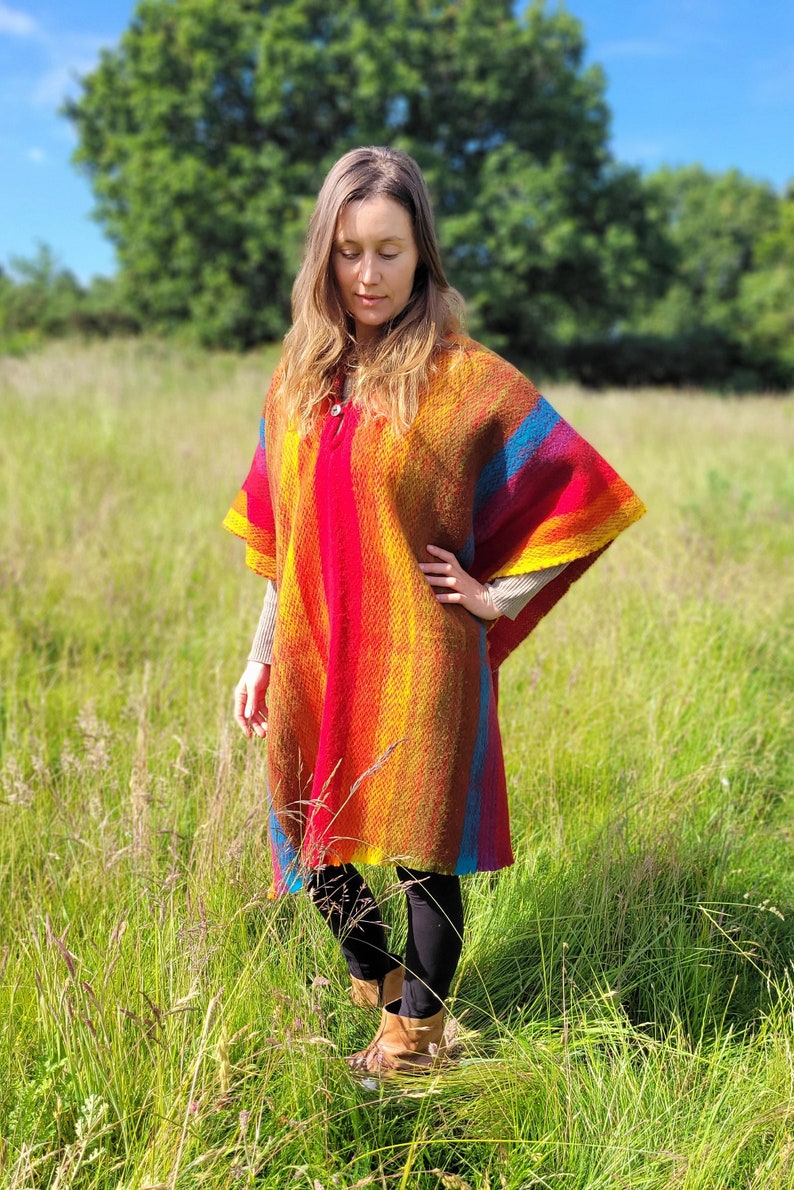 May include: A woman wearing a colorful striped poncho with a red, orange, yellow, and blue color scheme. The poncho is made of a soft, woven fabric and has a relaxed fit. The woman is standing in a field of tall grass.