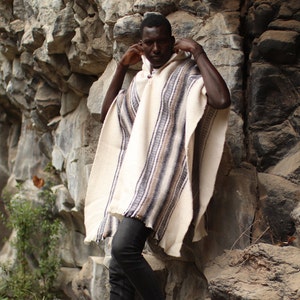 May include: A person wearing a white and brown striped poncho with a hood. The poncho is made of a soft, woven fabric. The person is standing in front of a rocky wall.