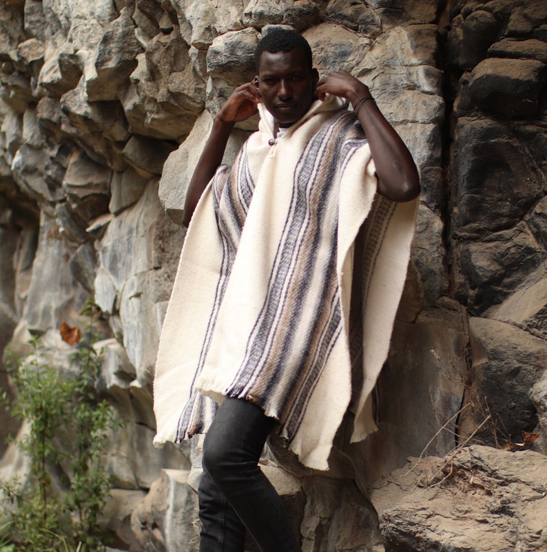May include: A person wearing a white and brown striped poncho with a hood. The poncho is made of a soft, woven fabric. The person is standing in front of a rocky wall.