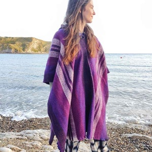 May include: A woman wearing a purple and white striped poncho with a hood. She is standing on a beach with the ocean in the background.