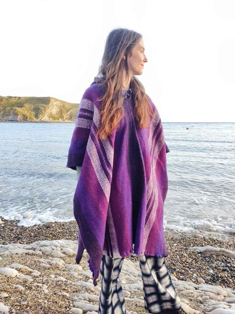 May include: A woman wearing a purple and white striped poncho with a hood. She is standing on a beach with the ocean in the background.