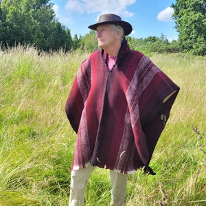 May include: A person wearing a red and black striped poncho with a brown hat standing in a field of tall grass.
