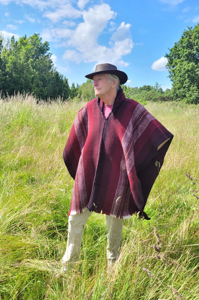 May include: A person wearing a red and black striped poncho with a brown hat standing in a field of tall grass.
