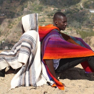 May include: Two people are sitting back-to-back on a sandy hill, wearing colorful striped ponchos with hoods. The person on the left is wearing a white and gray poncho, while the person on the right is wearing a red, orange, purple, and blue poncho.