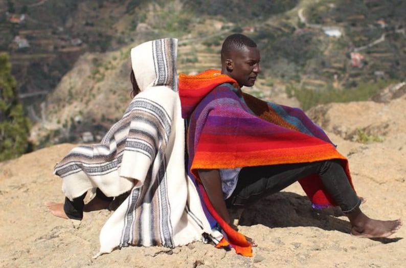 May include: Two people are sitting back-to-back on a sandy hill, wearing colorful striped ponchos with hoods. The person on the left is wearing a white and gray poncho, while the person on the right is wearing a red, orange, purple, and blue poncho.