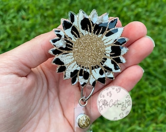 Puede incluir: Un girasol de estampado animal blanco y negro con purpurina dorada en el centro. El girasol está unido a un portagafete retráctil de plástico transparente con un botón de purpurina dorada. El portagafete tiene una correa de plástico transparente y un clip de metal plateado. El portagafete está etiquetado como "Cole's Customs 19".