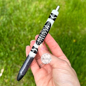 May include: A black and white crayon-shaped pen with the name "MELISSA" written on it. The pen has a black glitter finish on the bottom half.