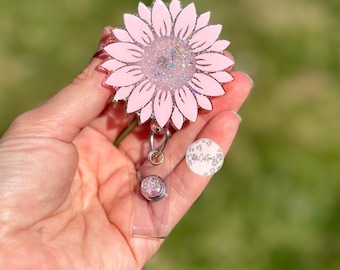 Puede incluir: Carrete de insignia de girasol rosa con purpurina, con un soporte de insignia de plástico transparente y un clip giratorio plateado. El girasol tiene un contorno blanco y un centro de purpurina plateado.