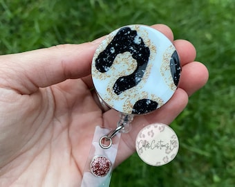 May include: A white, black, and gold leopard print retractable badge holder with a pink glitter accent. The badge holder has a clear plastic strap and a silver swivel clip. The text "Coda's Customs 19" is printed on the bottom of the badge holder.