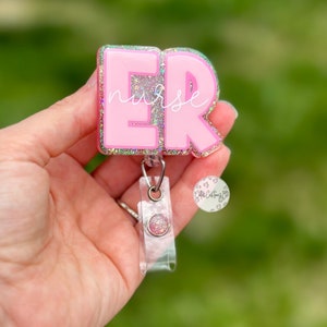 May include: Pink glitter badge holder with the letters "ER" and the word "nurse" written in white. The badge holder is attached to a clear retractable badge reel.