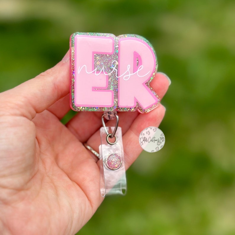 May include: Pink glitter badge holder with the letters "ER" and the word "nurse" written in white. The badge holder is attached to a clear retractable badge reel.