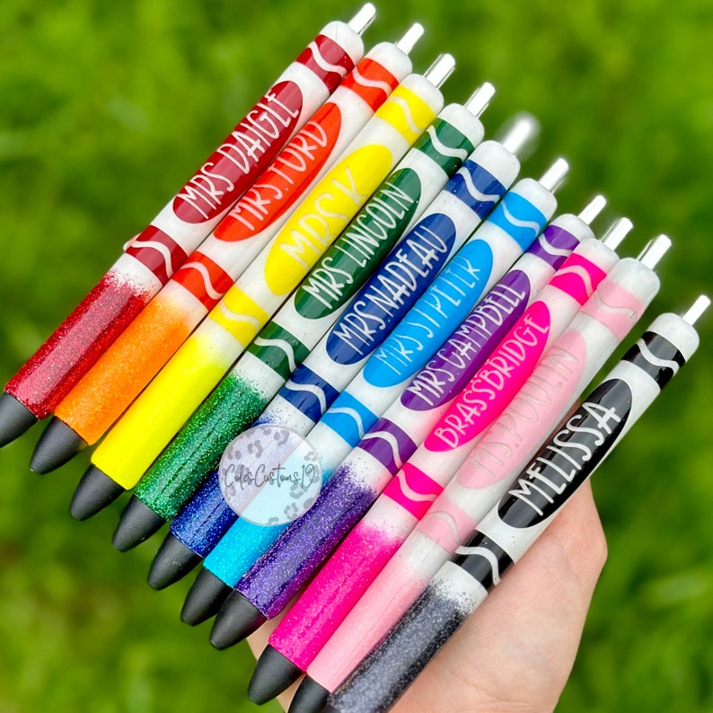 May include: A set of ten colorful glitter pens with white crayon-like barrels and black ink. Each pen has a different teacher's name written on it, including "Mrs. Daigle", "Mrs. Lincoln", and "Melissa".