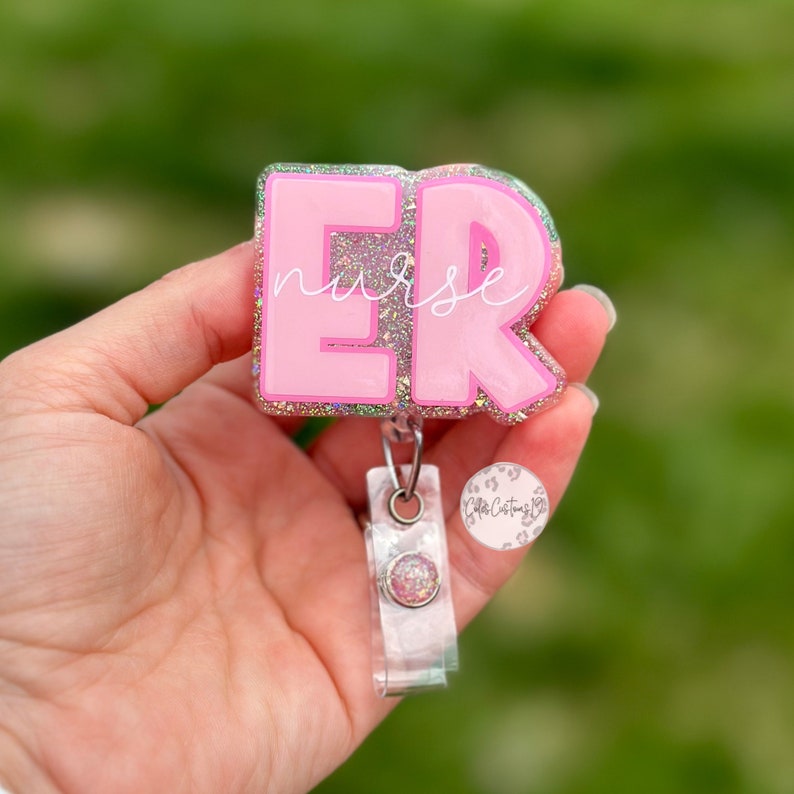 May include: Pink glitter resin badge holder with the letters "ER" in white and the word "nurse" written in white inside the "E". The badge holder is attached to a clear retractable badge reel with a pink glitter button.