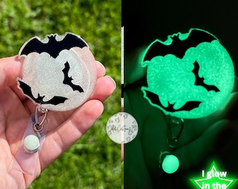 May include: A white and silver glitter badge reel with a black silhouette of three bats. The reel glows green in the dark and has the text "I glow in the dark" on a green star.