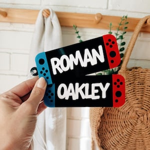 May include: Two rectangular luggage tags designed to resemble game controllers, featuring a black center with white text reading ROMAN and OAKLEY. Each tag has a blue left side and a red right side with black circular button details.
