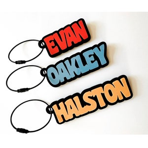 May include: Three colorful luggage tags with black keychains. The tags are made of a soft, flexible material and feature the names "Evan", "Oakley", and "Halston" in a bold, cartoon-style font.