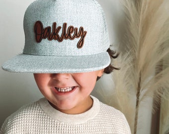 Puede incluir: Una gorra de camionero de malla gris y negra con un parche de cuero marrón que dice "Oakley" en letra cursiva.
