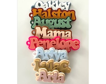 May include: A set of nine colorful, personalized name tags with a retro font style. The names are Oakley, Halston, August, Mama, Penelope, Ariva, Lilac, and Aria.