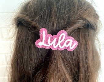 May include: Pink glitter hair clip with the name "Lula" written in white letters.
