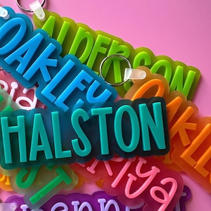 May include: A collection of colorful, translucent plastic keychains with names spelled out in bold letters. The names include Oakley, Halston, and Benni.