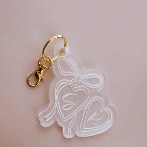 May include: A clear acrylic keychain with two hearts and a bow. The hearts have the letters "S" and "K" inside them.
