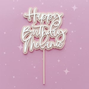 May include: A pink and white cake topper with the words "Happy Birthday Melanie" in a cursive font.