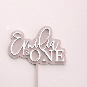 May include: Pink and white acrylic cake topper with the text "Emilia is ONE" in a cursive font.