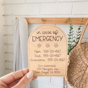 May include: A wooden sign with rounded edges and a light wood finish. The sign reads "in case of EMERGENCY" with three stylized flower icons. Below the text are contact numbers for Mom, Dad, and Grandma, followed by an address.