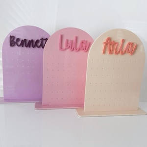 May include: Three pastel-coloured earring display stands. Each stand is arched at the top and has a name in a cursive font: "Bennett", "Lula", and "Aria". The stands are lavender, pink, and cream, with small holes for earrings.