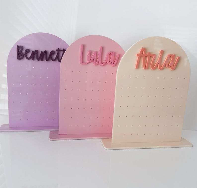 May include: Three pastel-coloured earring display stands. Each stand is arched at the top and has a name in a cursive font: "Bennett", "Lula", and "Aria". The stands are lavender, pink, and cream, with small holes for earrings.