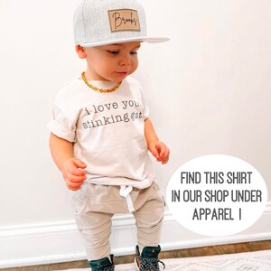 May include: A toddler wearing a light beige t-shirt with the text "i love you stinking gut" printed on it. The toddler is also wearing a gray and white baseball cap with a brown leather patch that says "Brooks" on it. The toddler is wearing beige pants and black and brown sneakers. The toddler is standing on a patterned rug.