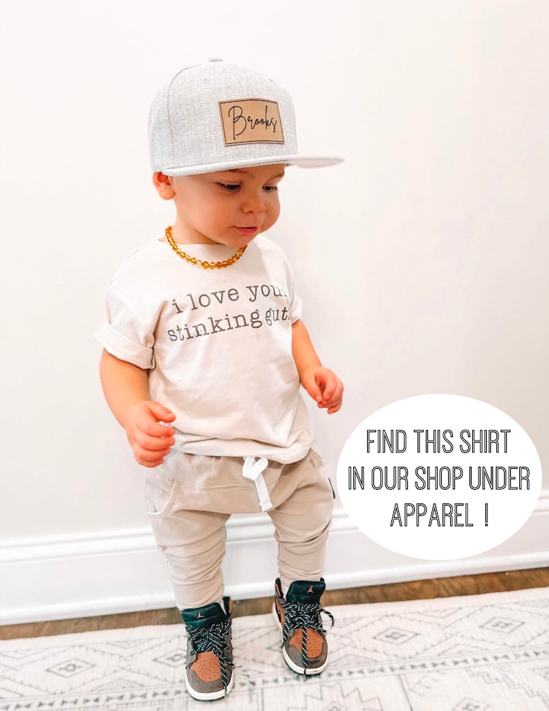 May include: A toddler wearing a light beige t-shirt with the text "i love you stinking gut" printed on it. The toddler is also wearing a gray and white baseball cap with a brown leather patch that says "Brooks" on it. The toddler is wearing beige pants and black and brown sneakers. The toddler is standing on a patterned rug.