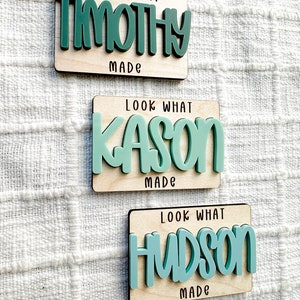 May include: Three wooden plaques with the text "Look What" and "Made" in black. Each plaque has a different name in teal: Timothy, Kason, and Hudson. The names are in a bold, playful font.