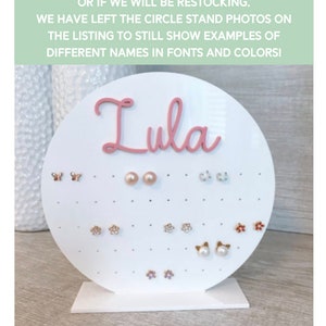May include: White circular earring display stand with a pink nameplate that reads "Lula". The stand has multiple holes for displaying earrings. The stand is filled with various earrings, including butterflies, flowers, and pearls.