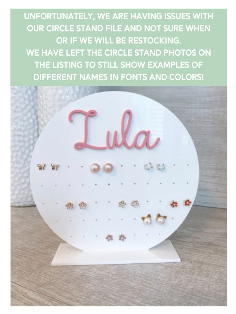 May include: White circular earring display stand with a pink nameplate that reads "Lula". The stand has multiple holes for displaying earrings. The stand is filled with various earrings, including butterflies, flowers, and pearls.