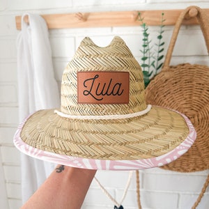 May include: A straw hat with a pink and white patterned brim and a brown leather patch that says "Lula" on the front.