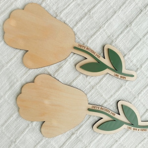 K&ouml;nnte beinhalten: Zwei Holzdekorationen in Tulpenform. Die Tulpenk&ouml;pfe sind hellbraun, mit gr&uuml;nen Bl&auml;ttern und Stielen. Auf den Stielen steht der Text "HAPPY MOTHERS DAY" und "LOVE 4ever & always". Die Dekorationen befinden sich auf einer wei&szlig;en, strukturierten Oberfl&auml;che.