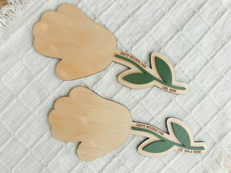 K&ouml;nnte beinhalten: Zwei Holzdekorationen in Tulpenform. Die Tulpenk&ouml;pfe sind hellbraun, mit gr&uuml;nen Bl&auml;ttern und Stielen. Auf den Stielen steht der Text "HAPPY MOTHERS DAY" und "LOVE 4ever & always". Die Dekorationen befinden sich auf einer wei&szlig;en, strukturierten Oberfl&auml;che.